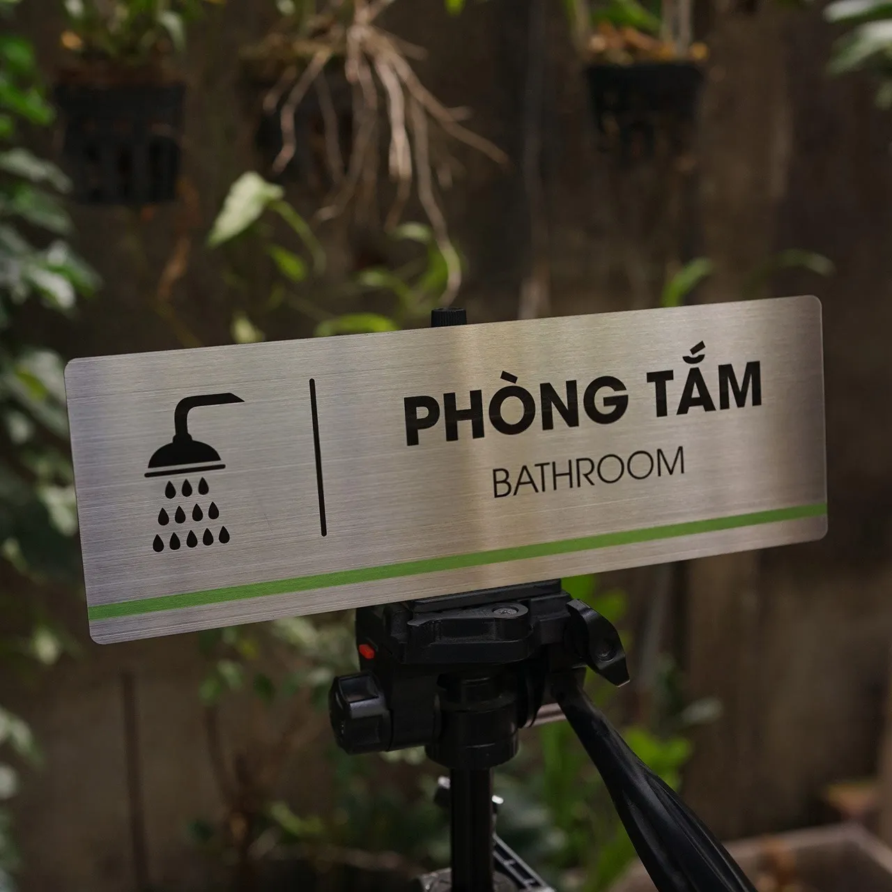 Bảng tên phòng tắm inox 304 – góc chụp thấp, thấy rõ hiệu ứng bề mặt phay xước Bảng tên phòng tắm inox 304 phay xước góc chụp thấp trên tripod nền tường gỗ