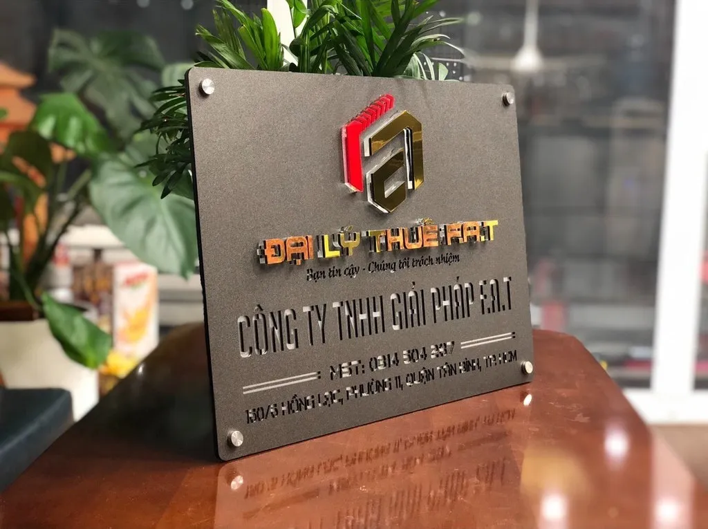 Bảng hiệu công ty inox xám khói logo nổi 3D phối cảnh đặt trên bàn văn phòng Bảng hiệu công ty inox xám khói logo 3D vàng đỏ bạc phối cảnh thực tế đặt trên bàn văn phòng cây xanh phía sau