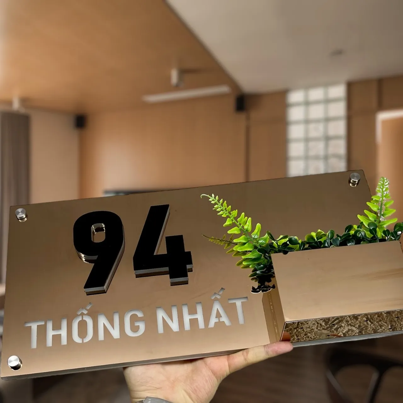 Bảng số nhà inox vàng gương — góc cao nhìn xuống thấy chậu cây tích hợp và mặt gương phản chiếu Bảng số nhà inox vàng gương tích hợp chậu cây góc cao nhìn xuống thấy đất trồng cây trong chậu