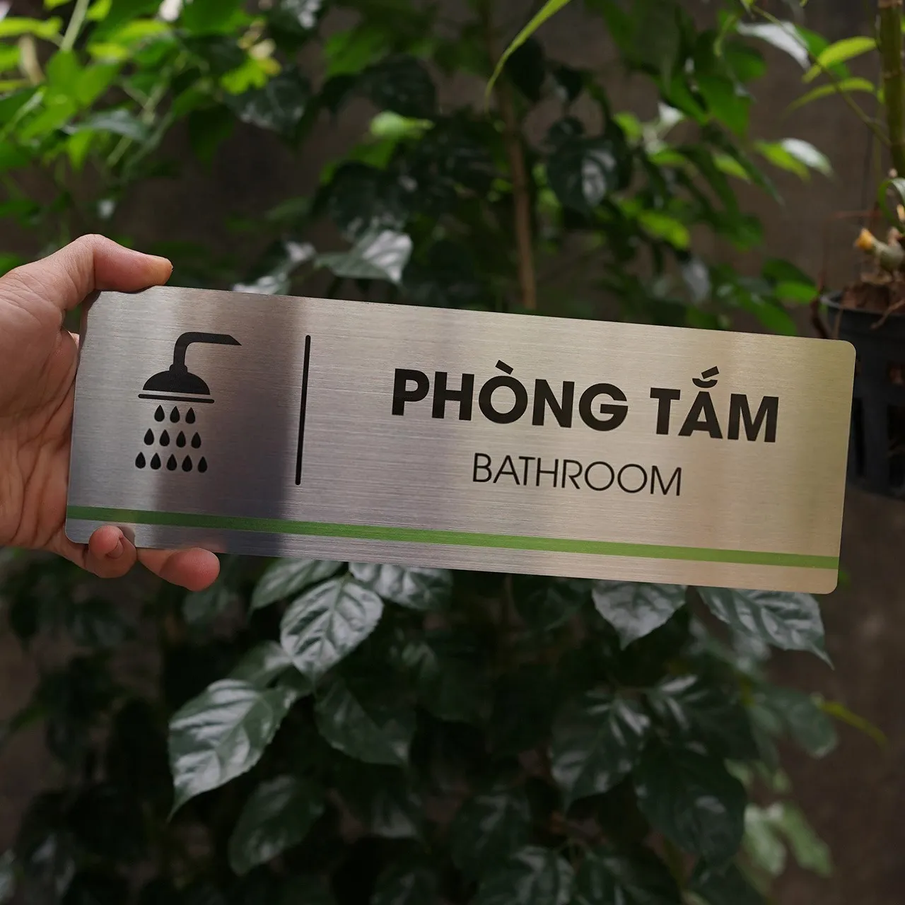 Cầm tay bảng tên phòng tắm inox 304 – mặt trước nền cây xanh Bảng tên phòng tắm inox 304 phay xước cầm tay mặt trước nền cây xanh sáng