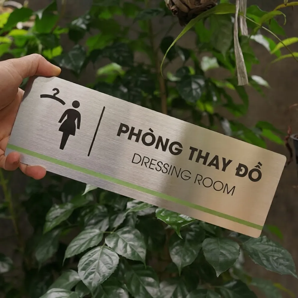 Cầm tay bảng tên phòng thay đồ inox 304 trắng xước mặt trước thấy rõ bộ đôi pictogram móc áo và nhân vật nữ