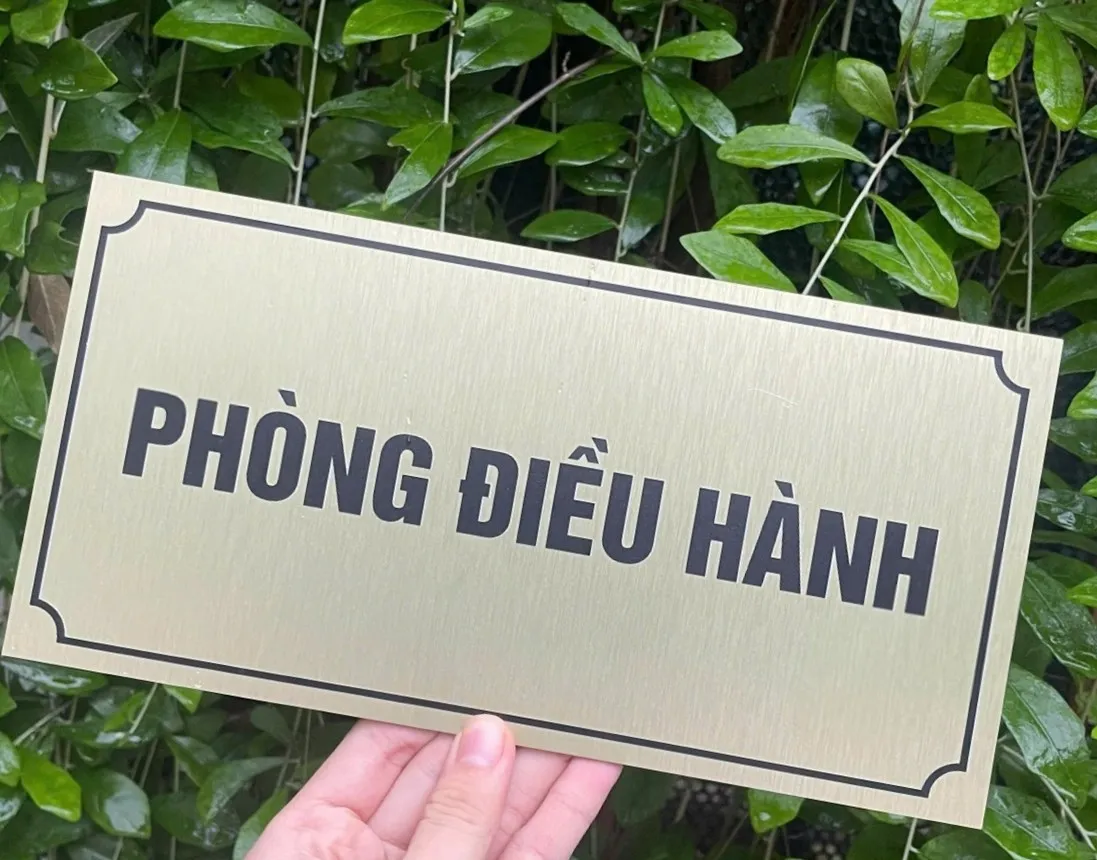 Bảng tên phòng điều hành inox vàng xước – chi tiết viền khung Bảng tên phòng điều hành inox vàng xước cầm tay nền lá xanh
