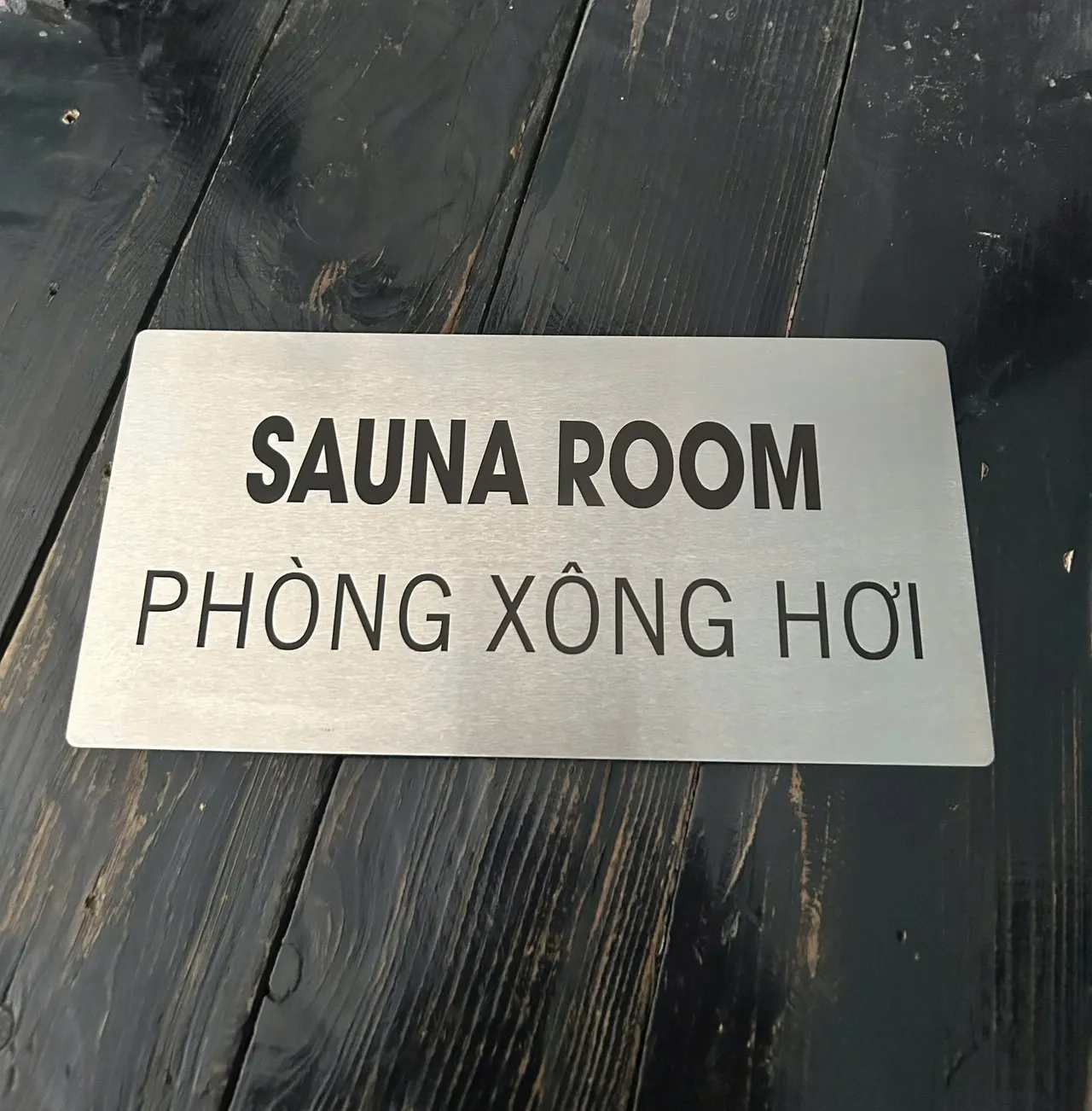 Biển Sauna Room - Phòng Xông Hơi inox bạc xước Biển Sauna Room - Phòng Xông Hơi inox bạc xước song ngữ Anh Việt trên nền gỗ
